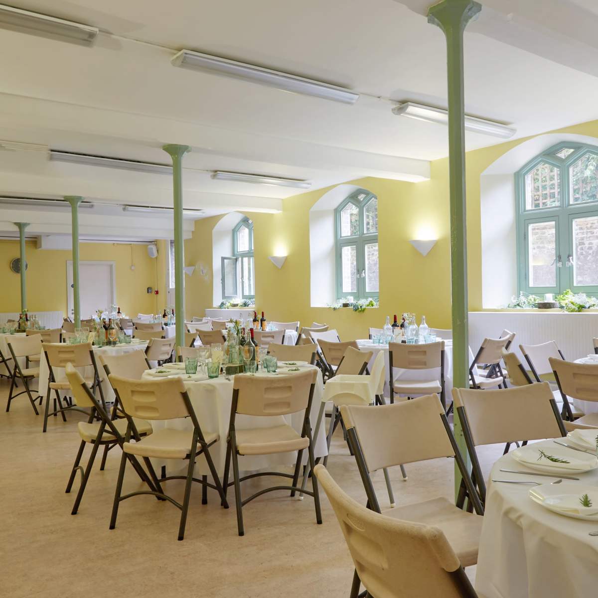 A spacious dining area features numerous tables set for a meal, with green windows and light-colored walls.