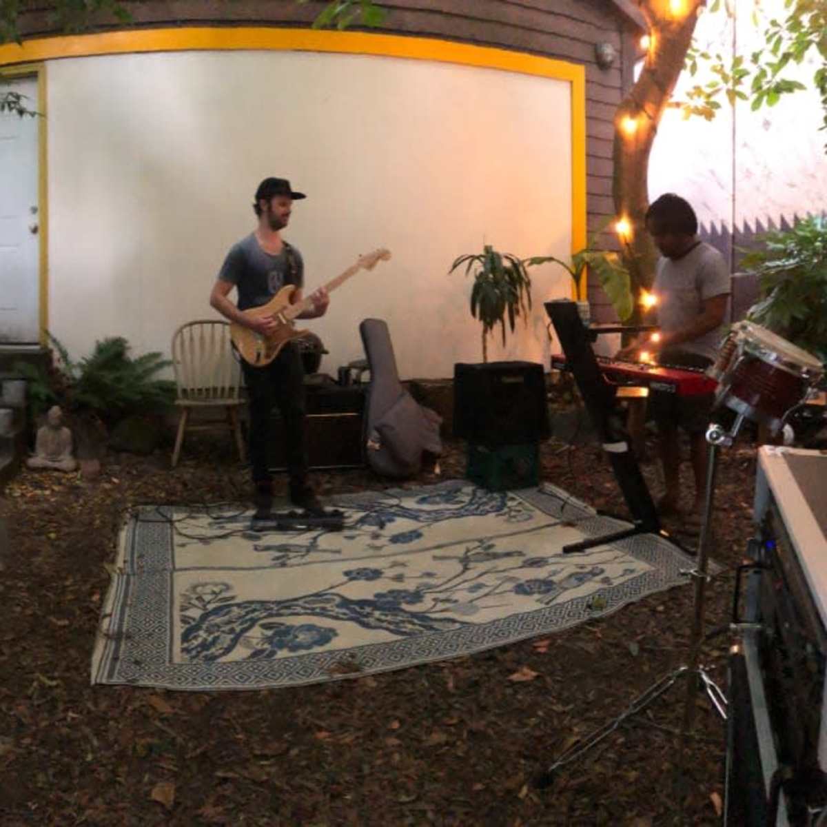 Three musicians perform in a garden space, with one on drums, one on guitar, and one on keyboards, surrounded by plants and decorative lighting.