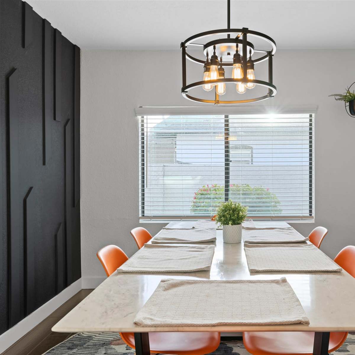 A modern dining area features a long table with orange chairs, a pendant light, and a window with blinds allowing natural light to enter.