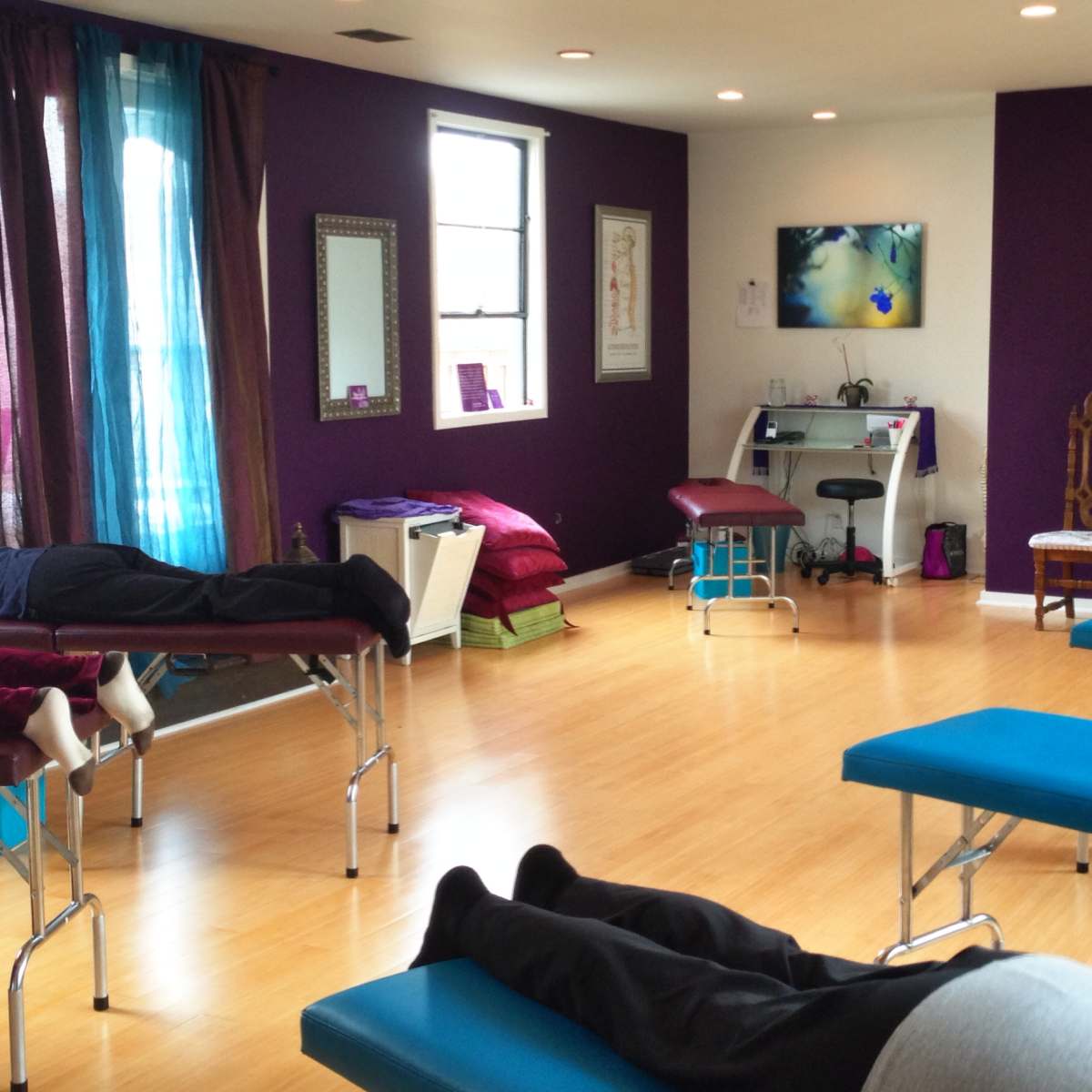 A brightly lit therapy room with several treatment tables occupied by individuals lying down, surrounded by purple walls and calming decor.