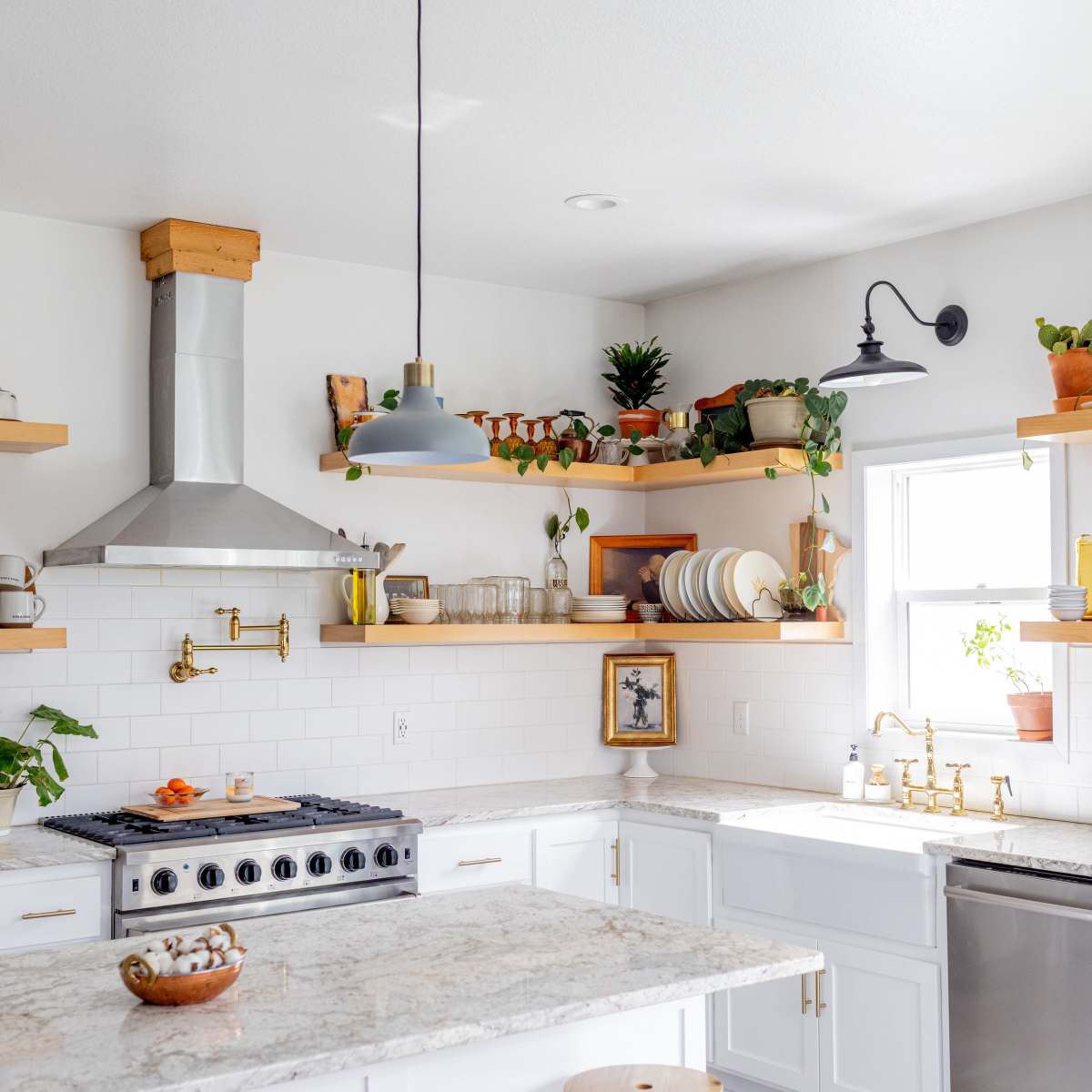 A modern kitchen features white cabinetry, open shelving with plants and dishware, and a large countertop with a stainless steel stove.