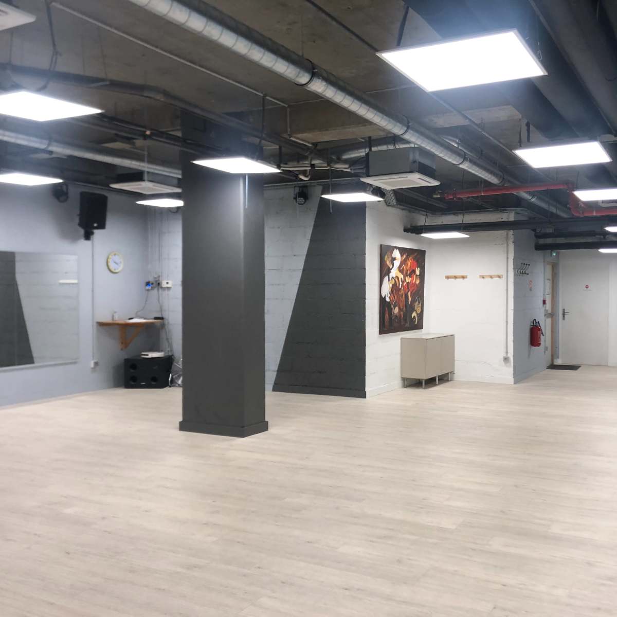 An empty, well-lit studio space featuring a wooden floor, a wall mirror, and a large artwork on one wall.