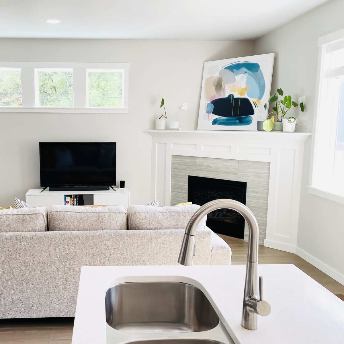 A light-filled living room features a beige sofa facing a television on a white media console, with a modern fireplace and artwork on the wall.