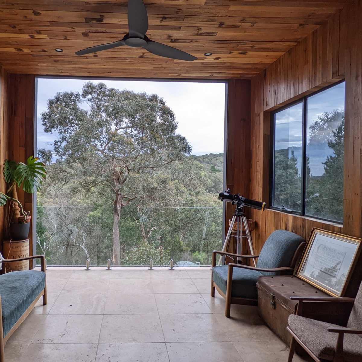 A wooden-paneled room with large windows overlooking a natural landscape, featuring two couches, a telescope, and a few pieces of furniture.
