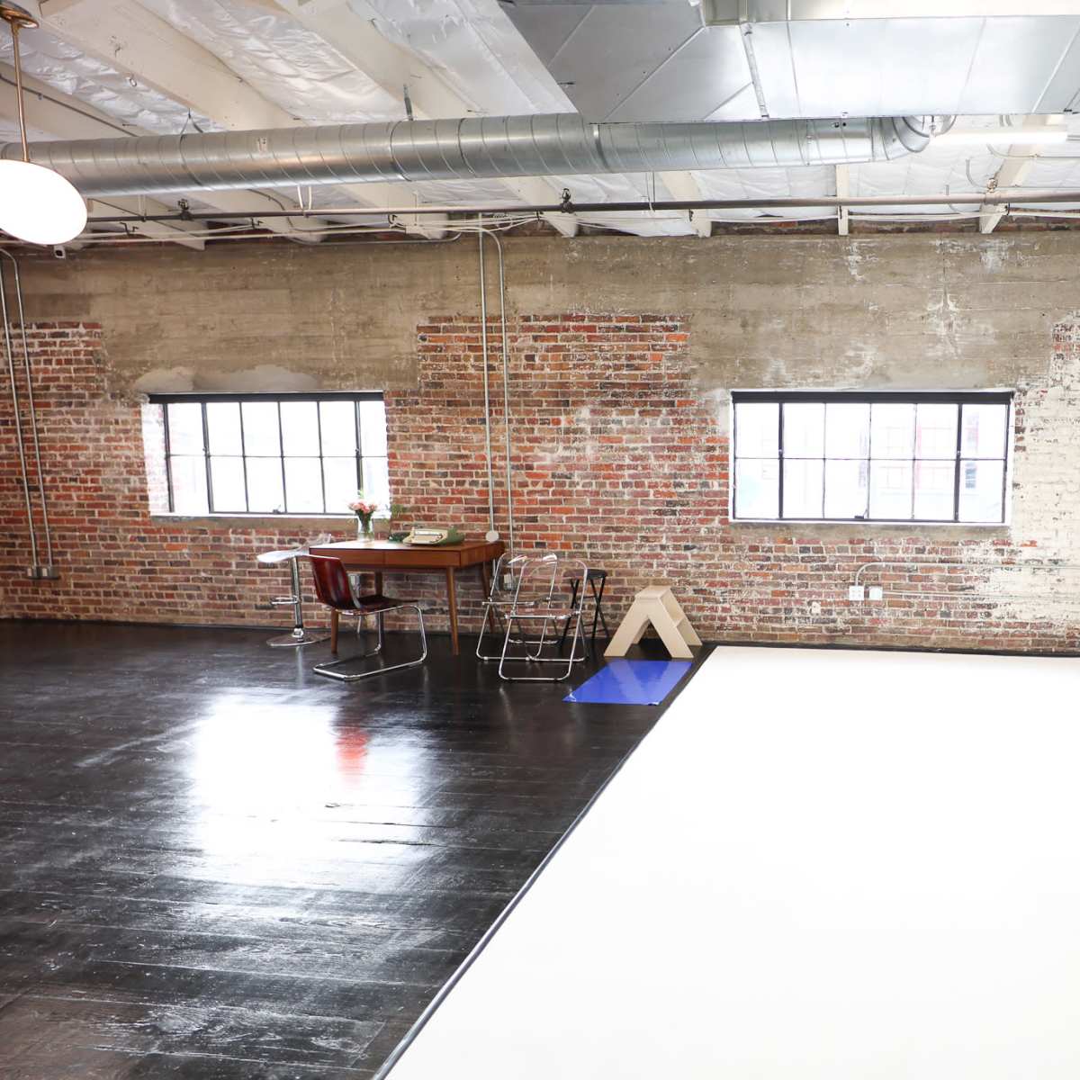 A spacious studio featuring exposed brick walls, large windows, and a clean white backdrop, with a dining table and chairs set off to the side.
