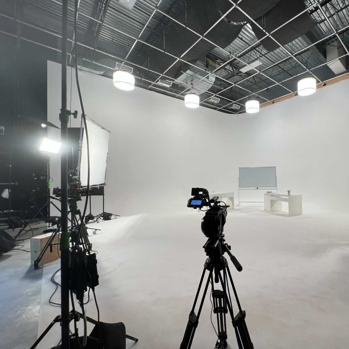 A film studio setup features a white backdrop, two cameras on tripods, a whiteboard, and several lighting fixtures.