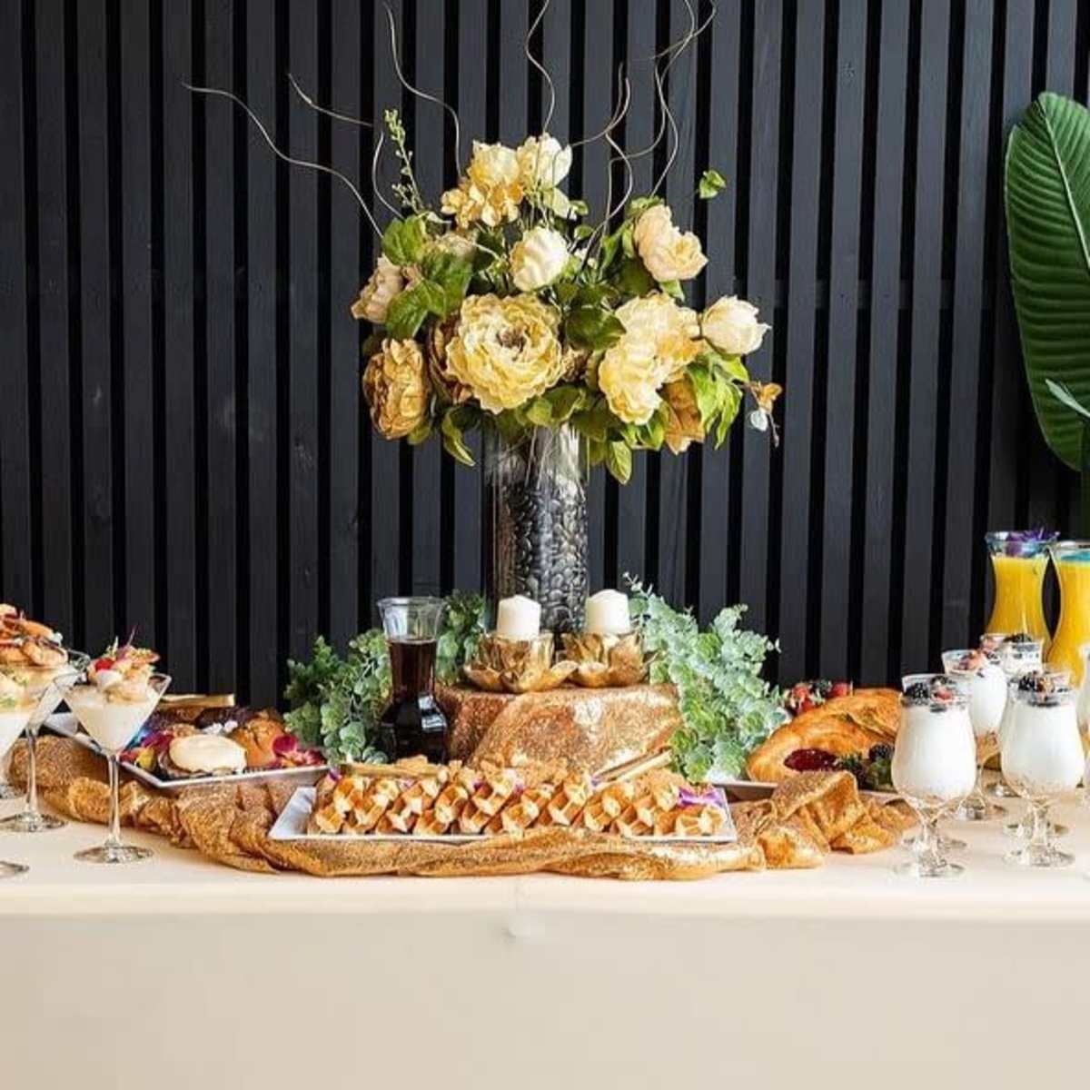 A beautifully arranged buffet table features a floral centerpiece, an assortment of desserts, baked goods, and beverages.