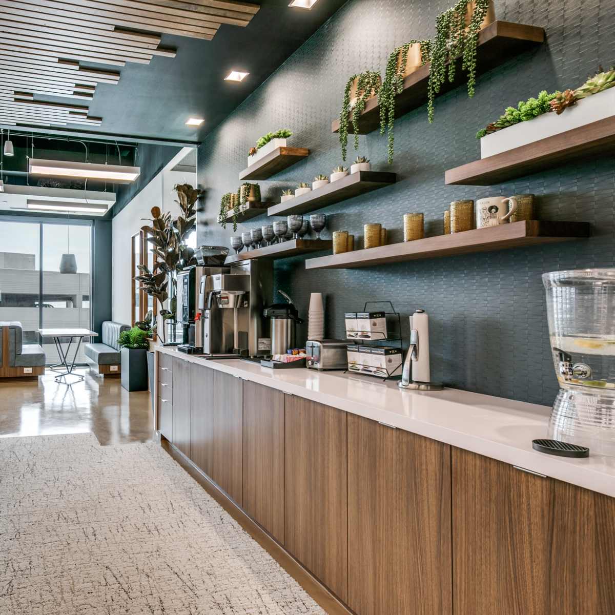 A modern kitchen area with wooden cabinetry, a countertop, a coffee station, and a water dispenser, surrounded by large windows that provide natural light.