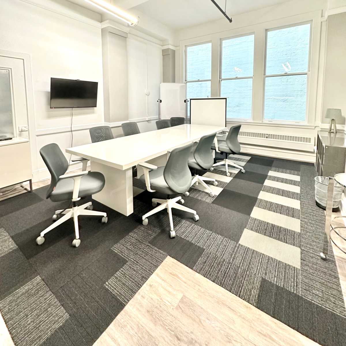 A modern conference room with a long white table, eight gray chairs, large windows, and a television mounted on the wall.