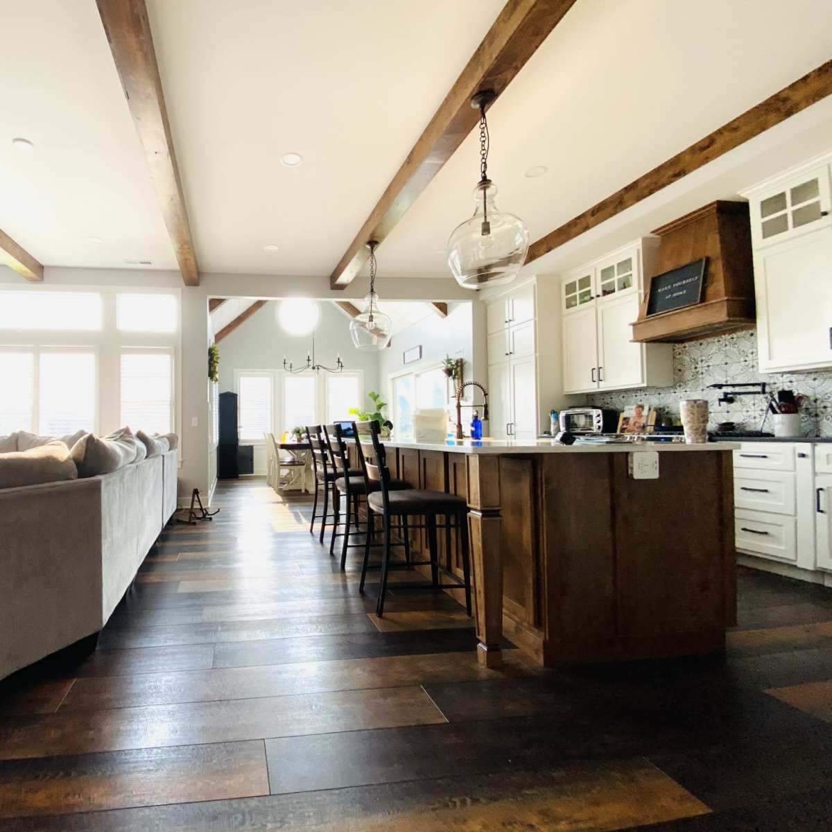 A modern kitchen and living area featuring wooden beams, a large sofa, a dining table with chairs, and ample natural light from several windows.