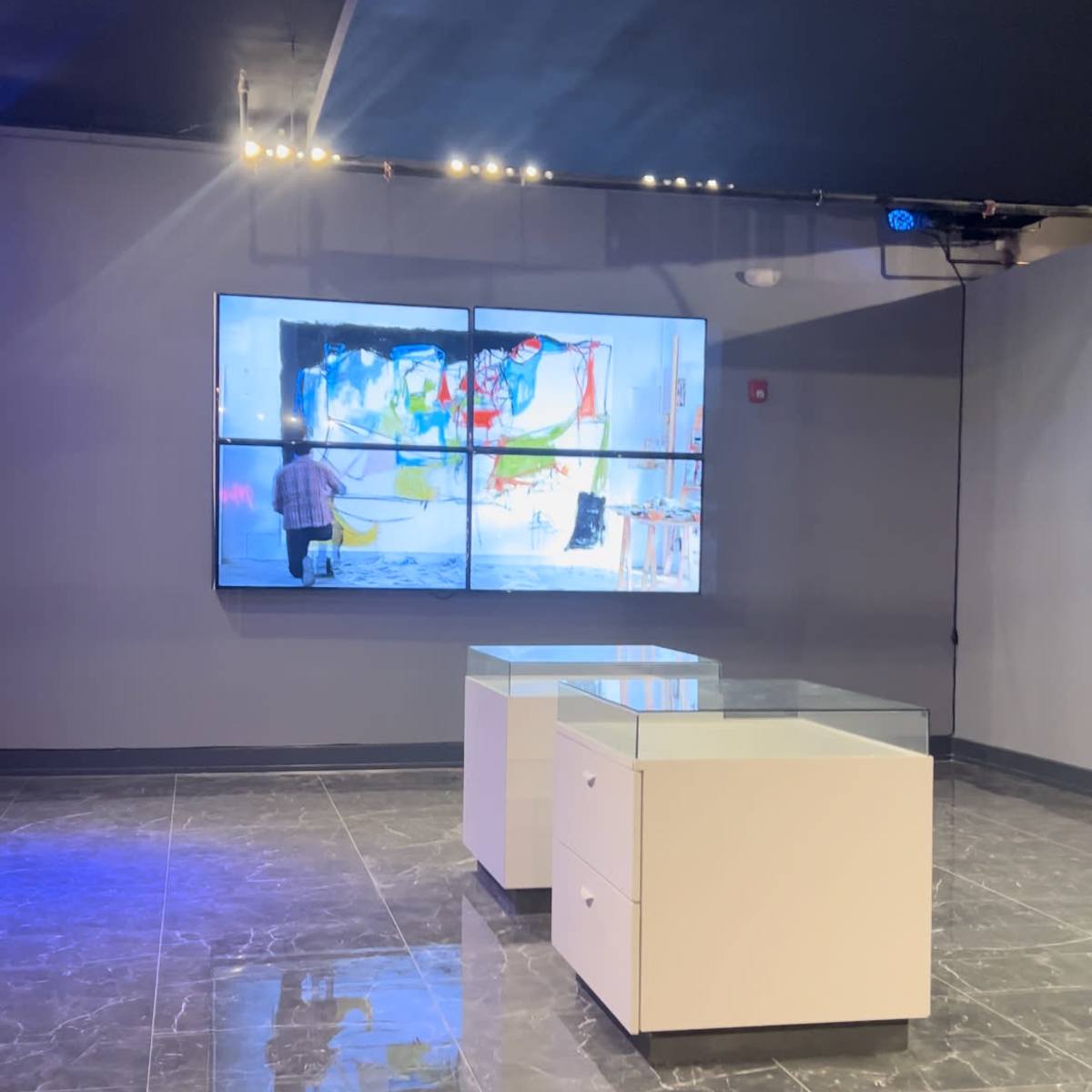 A modern gallery space featuring a large display screen showing an artist at work, with two white display tables in the foreground.