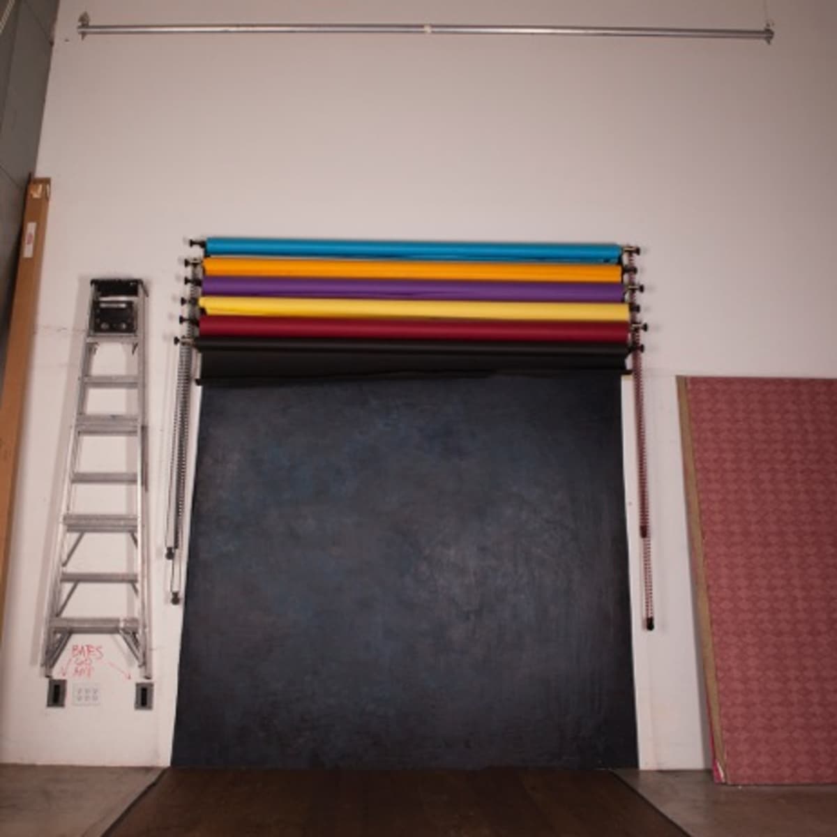 A photography studio setup with a large black backdrop, a ladder, and several colored backdrops hanging from a rod on the wall.