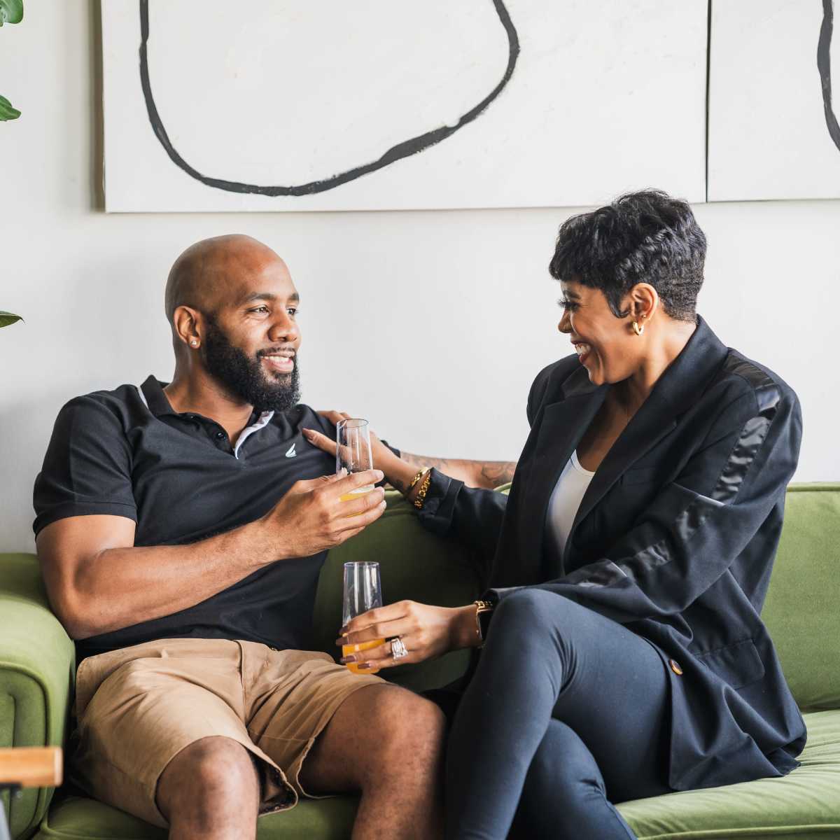 A man and woman sit together on a green sofa, laughing and enjoying drinks in a stylish living room.