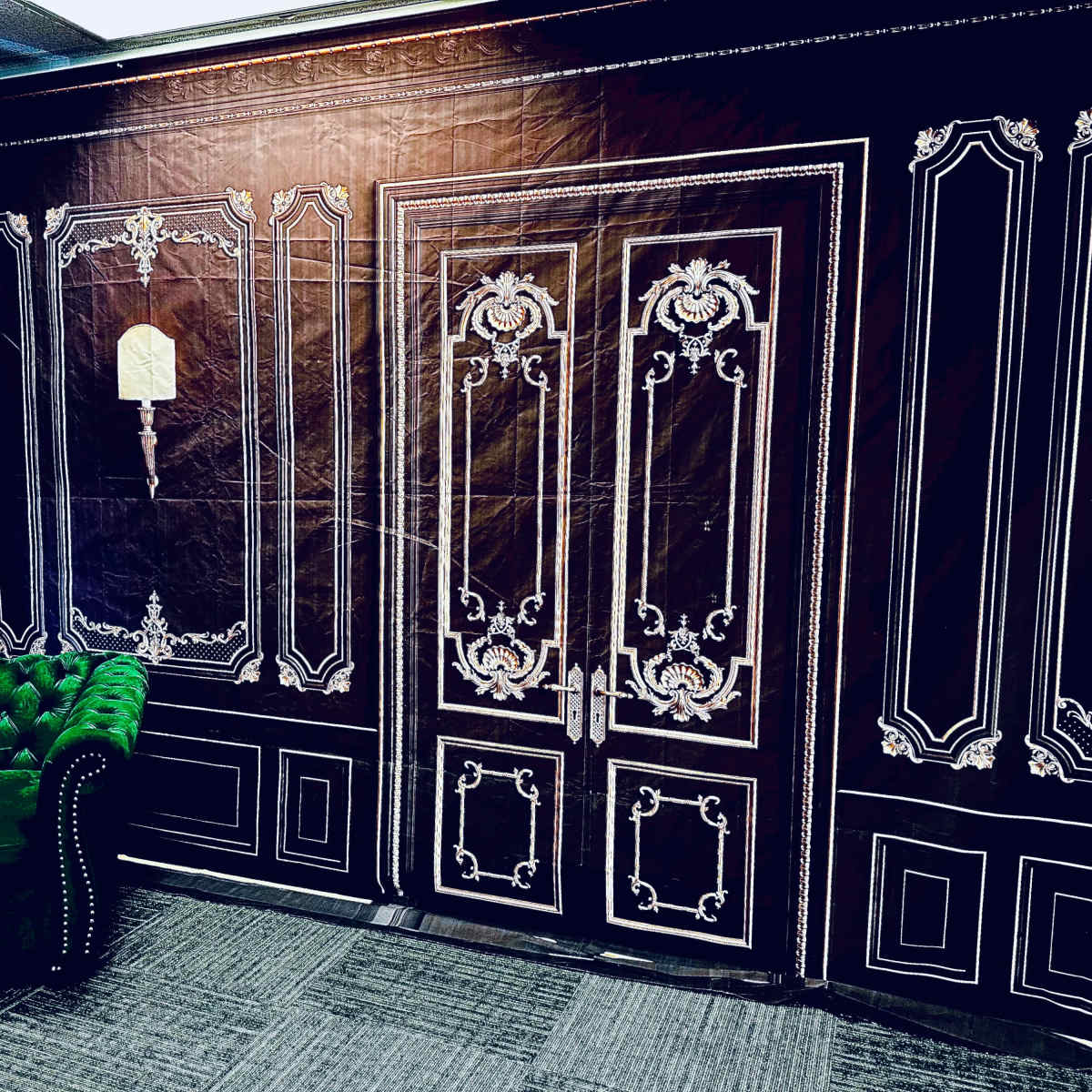 A room with a black and white ornate wallpaper featuring a door design, alongside a green velvet couch.