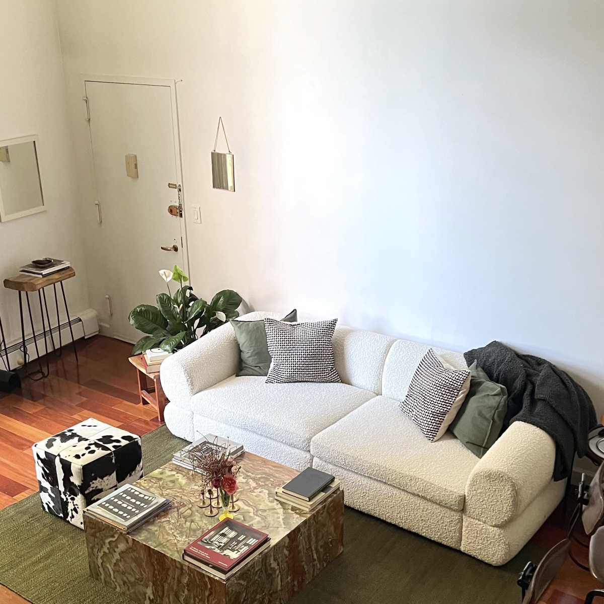 The room features a white, textured couch, a patterned coffee table, and a green area rug, with wooden flooring and a few plants adding to the decor.