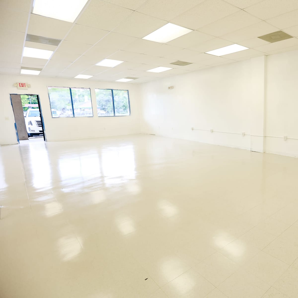 A large, empty room with white walls and a glossy floor, illuminated by overhead fluorescent lights and featuring several windows.