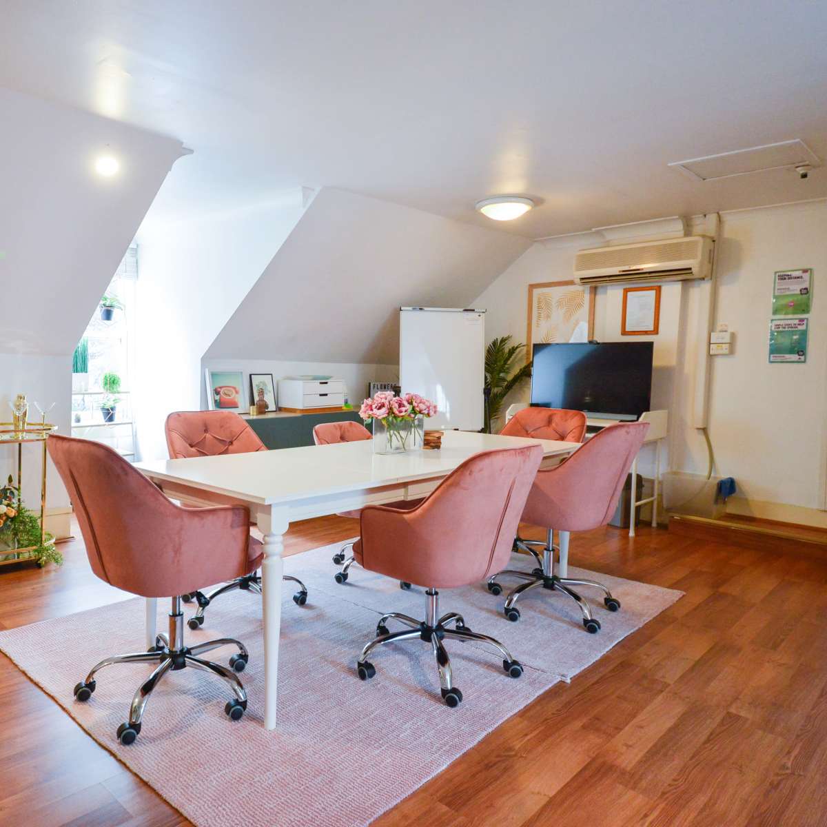 A modern meeting room with a white table surrounded by pink chairs, a notice board, and decorative plants.