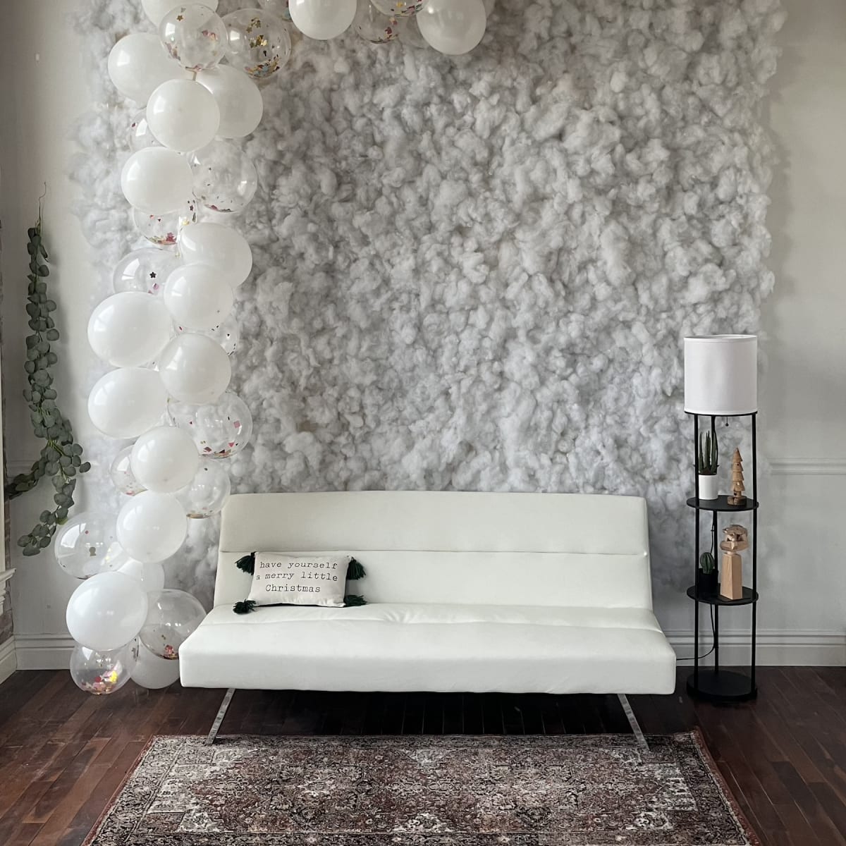 A white sofa is positioned in front of a textured gray wall adorned with white balloons and a decorative lamp on a black stand.