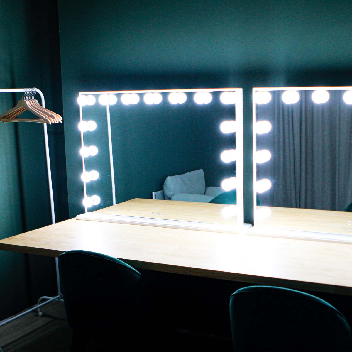 A well-lit vanity area features two mirrors surrounded by bright bulbs, set against a dark green wall, with a wooden table and a clothing hanger nearby.