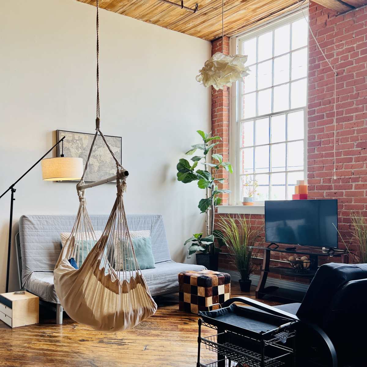 A cozy living space featuring a hammock chair, a gray sofa bed, a small television on a stand, and plants against exposed brick walls.
