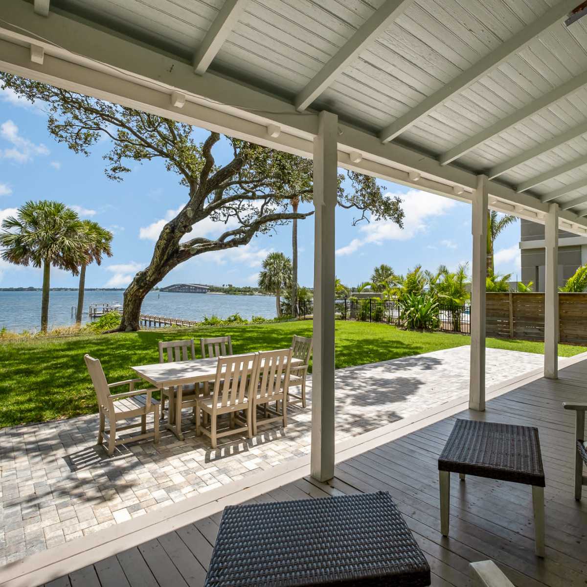 A covered patio with wooden furniture overlooks a grassy area and a waterfront scene.