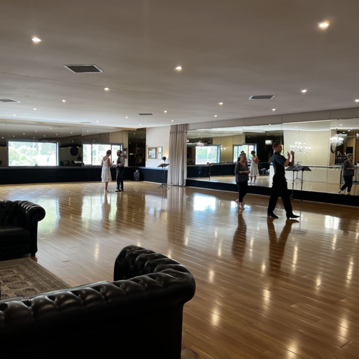 A spacious dance studio features polished wooden floors, large mirrors, and couples practicing dancing while others observe.