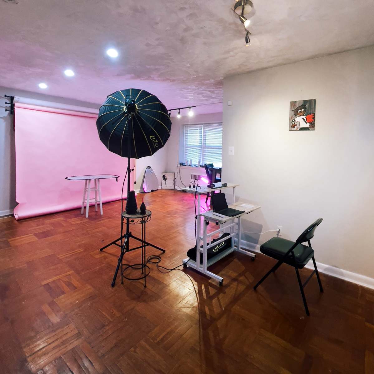 A bright studio space features a pink backdrop, a photography umbrella light, and a small desk setup.