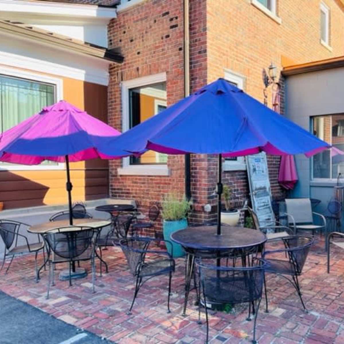 An outdoor patio features several round tables and chairs, shaded by large purple umbrellas, next to a brick wall and a gray building.