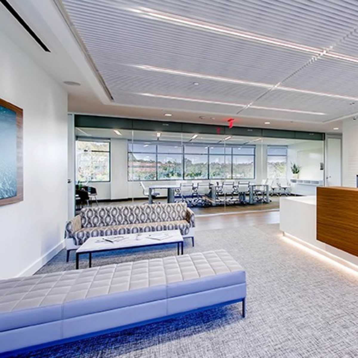 A modern office reception area featuring a wooden front desk, seating arrangements with sofas, and a glass-walled conference room in the background.