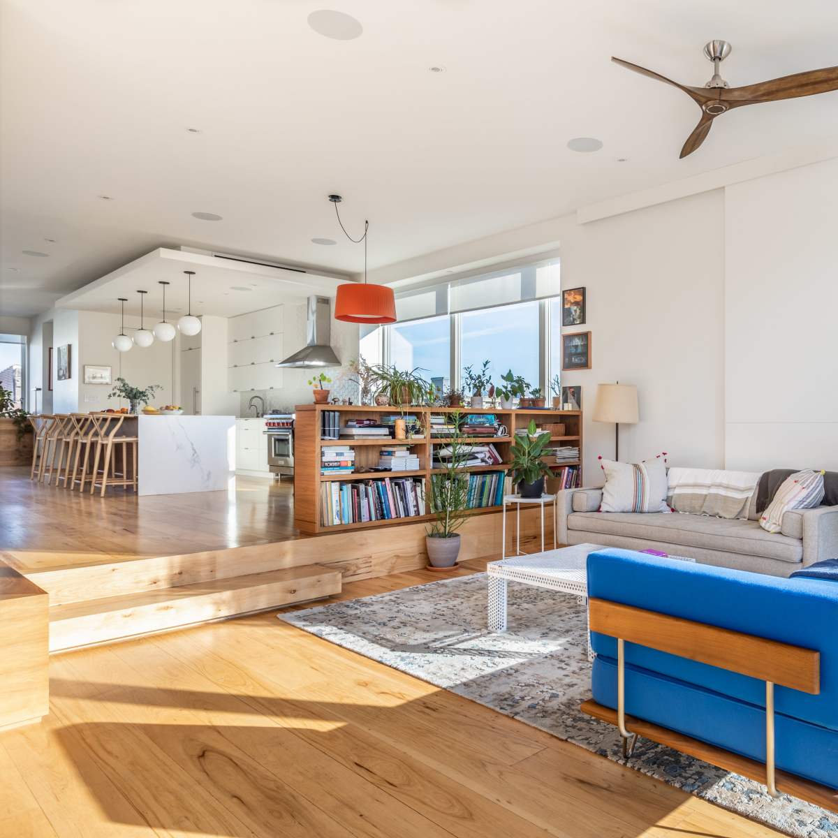 A bright and modern living room featuring a blue sofa, a coffee table, and a bookshelf, with an open kitchen visible in the background.