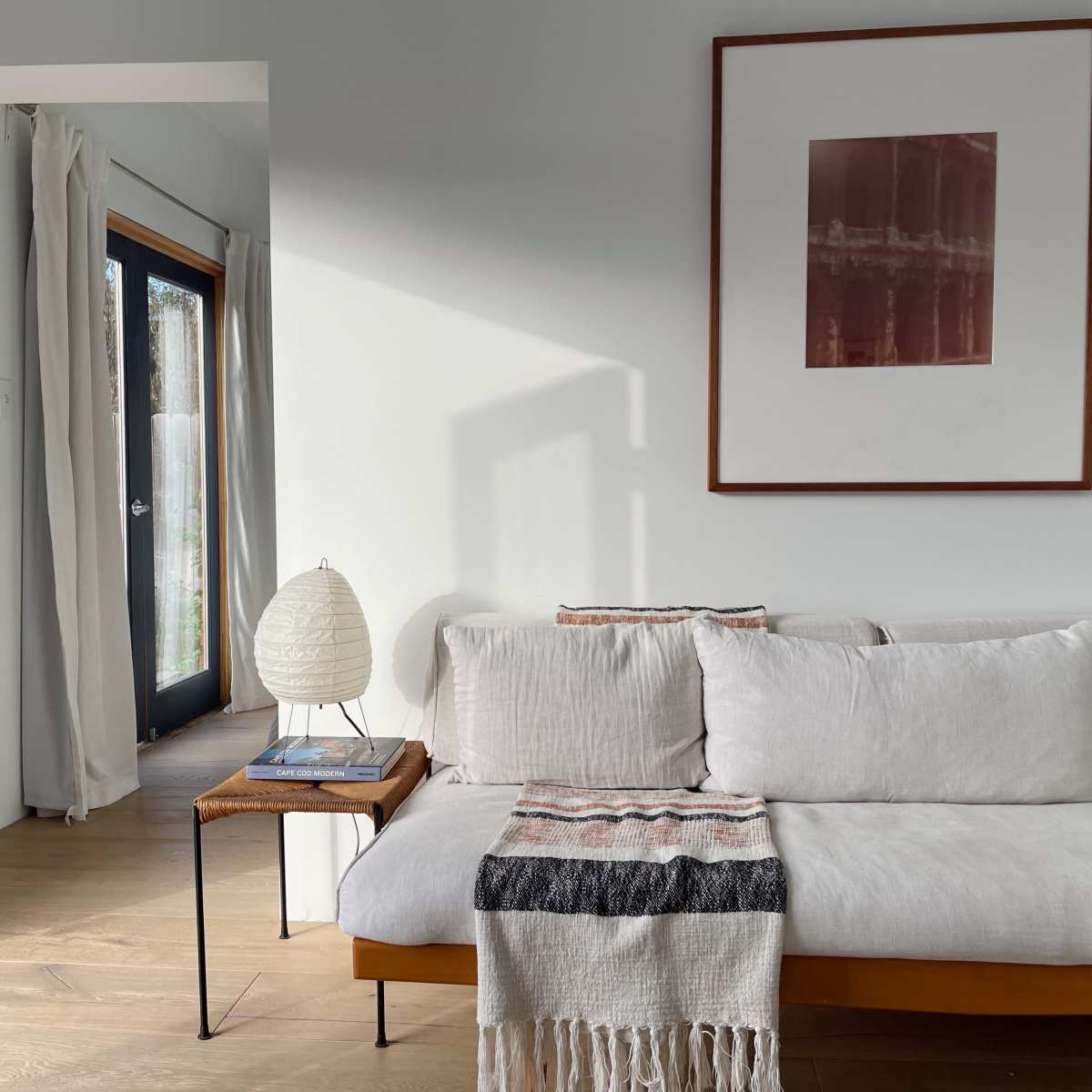 A light-filled living space features a sofa with neutral cushions, a small wooden side table, and a framed artwork on the wall.