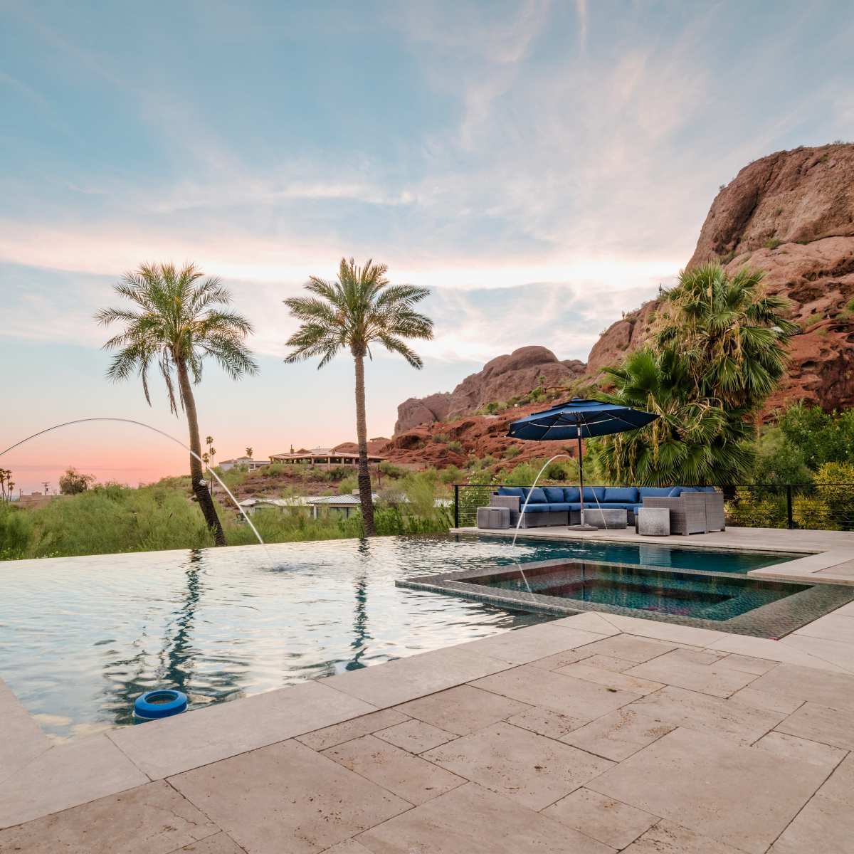 A swimming pool with an attached spa, surrounded by palm trees and set against a backdrop of rocky hills under a colorful sunset sky.