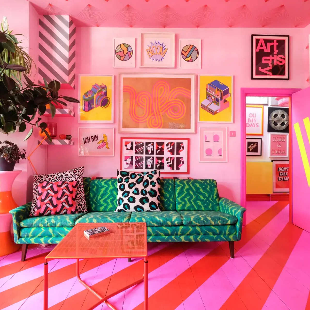 The room features bright pink walls, a green couch with patterned cushions, and a variety of colorful artwork and decor.