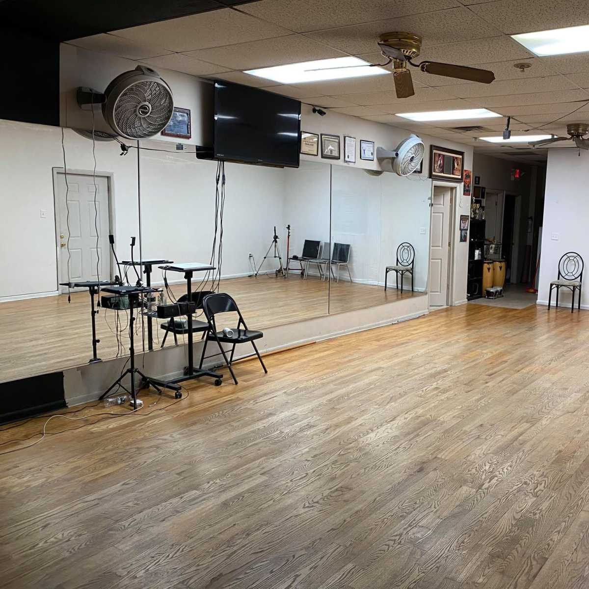 A spacious dance studio with wooden floors, a large mirror along one wall, and several black chairs arranged on the side.