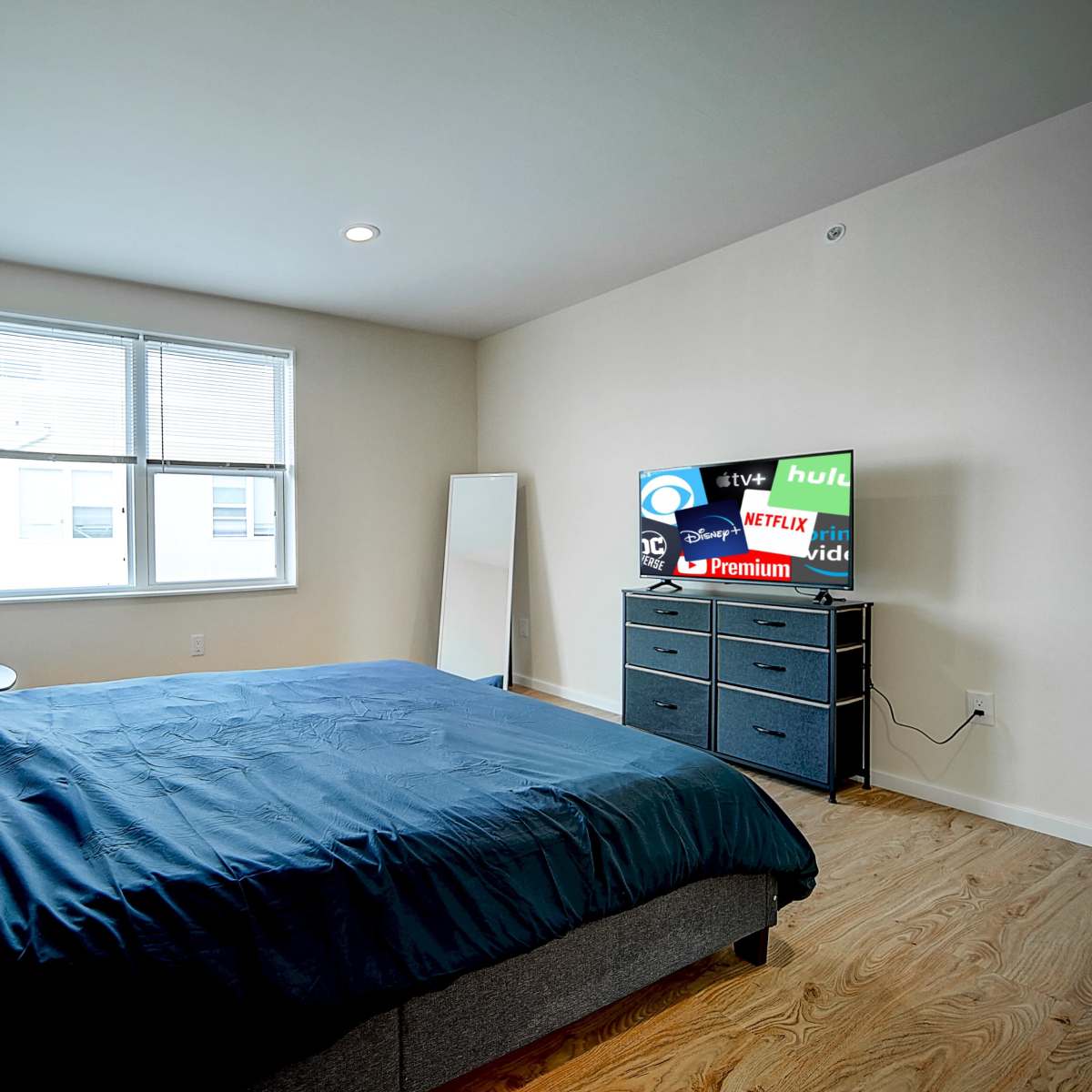 A bedroom with a large bed, a mirror, and a dresser featuring a television displaying various streaming service logos.