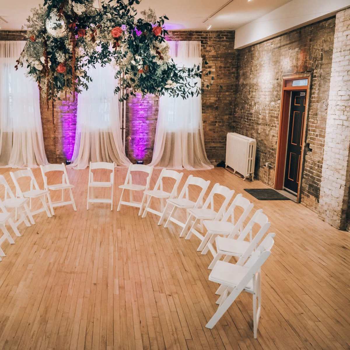 A circular arrangement of white chairs in an indoor space, with draped fabric and floral decorations above.