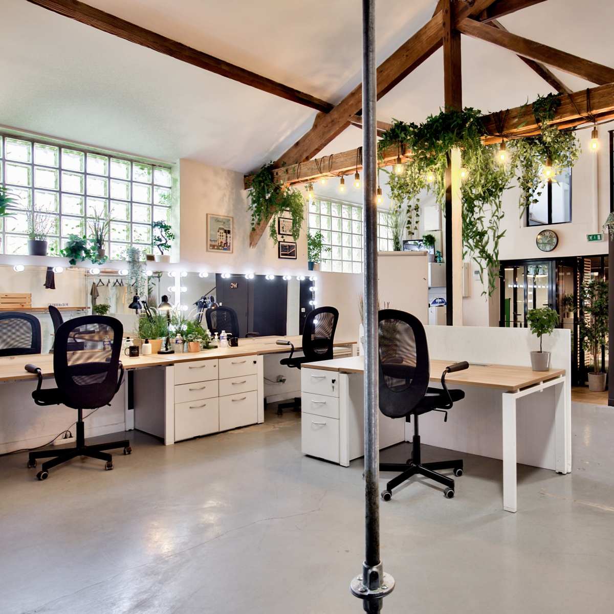 A spacious office interior featuring multiple workstations with plants, large windows, and an adjacent staircase.