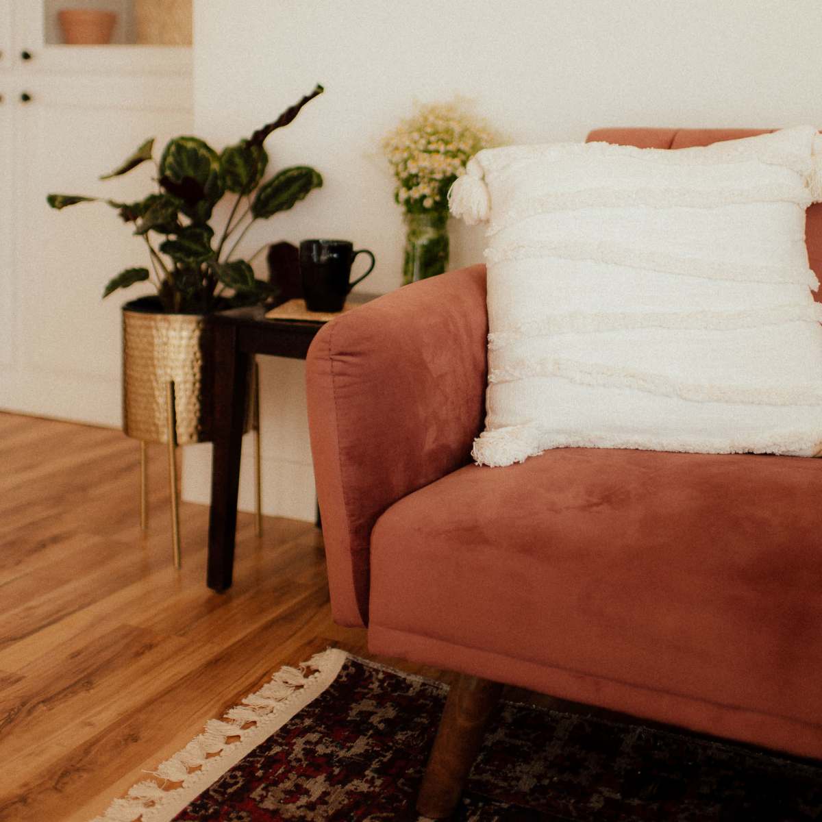 A pink sofa with a textured white pillow sits beside a small wooden table, accented by a plant and a black cup, in a bright room with wood flooring.