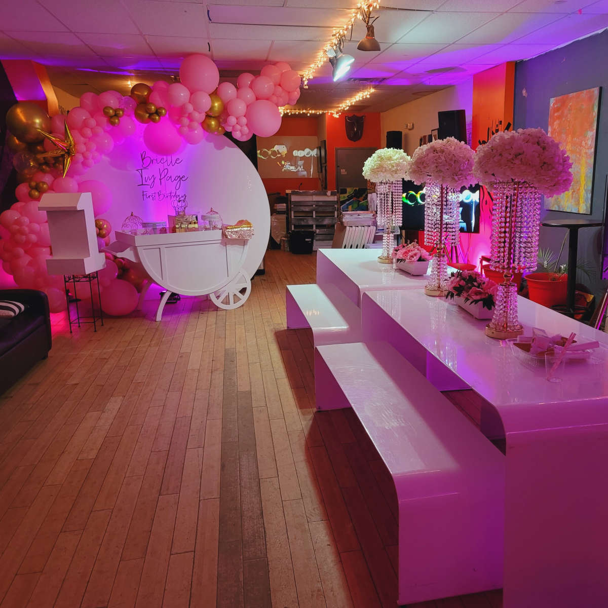 A brightly lit party venue with a decorative setup featuring tables, floral centerpieces, and balloon arches in a variety of colors.