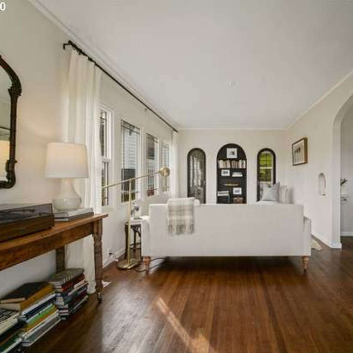 A bright living room with a white couch, a wooden side table, and a bookshelf against the wall, illuminated by natural light from multiple windows.