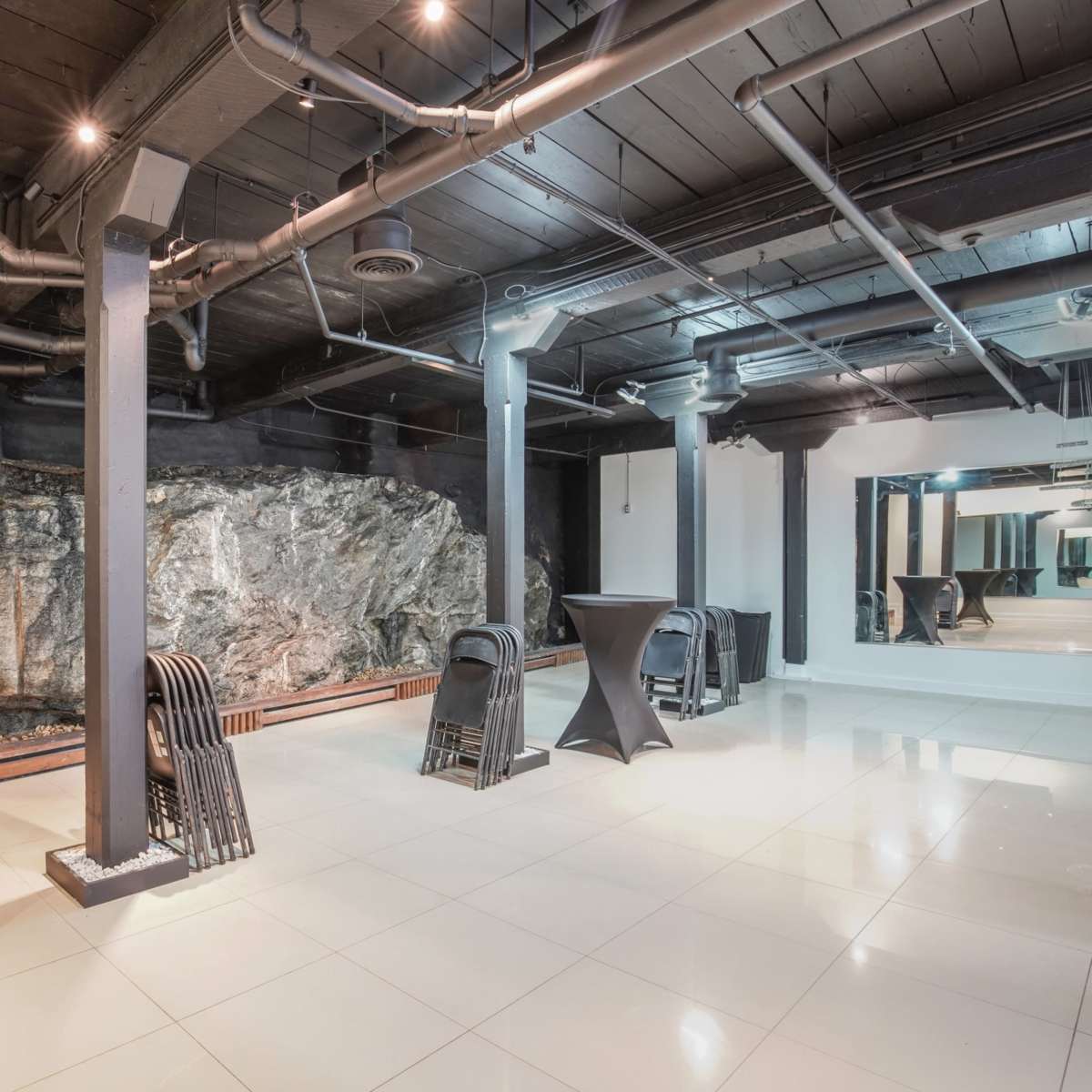An industrial-style indoor space featuring a large rock wall, black columns, and several black cocktail tables set on a tiled floor.