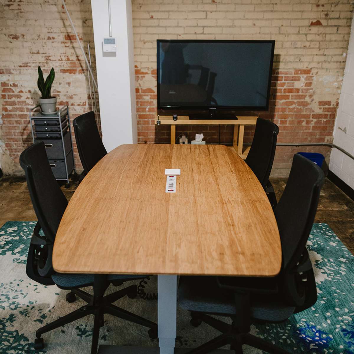 A rectangular wooden conference table with four black chairs is centered in a room featuring exposed brick walls and a television on a stand.