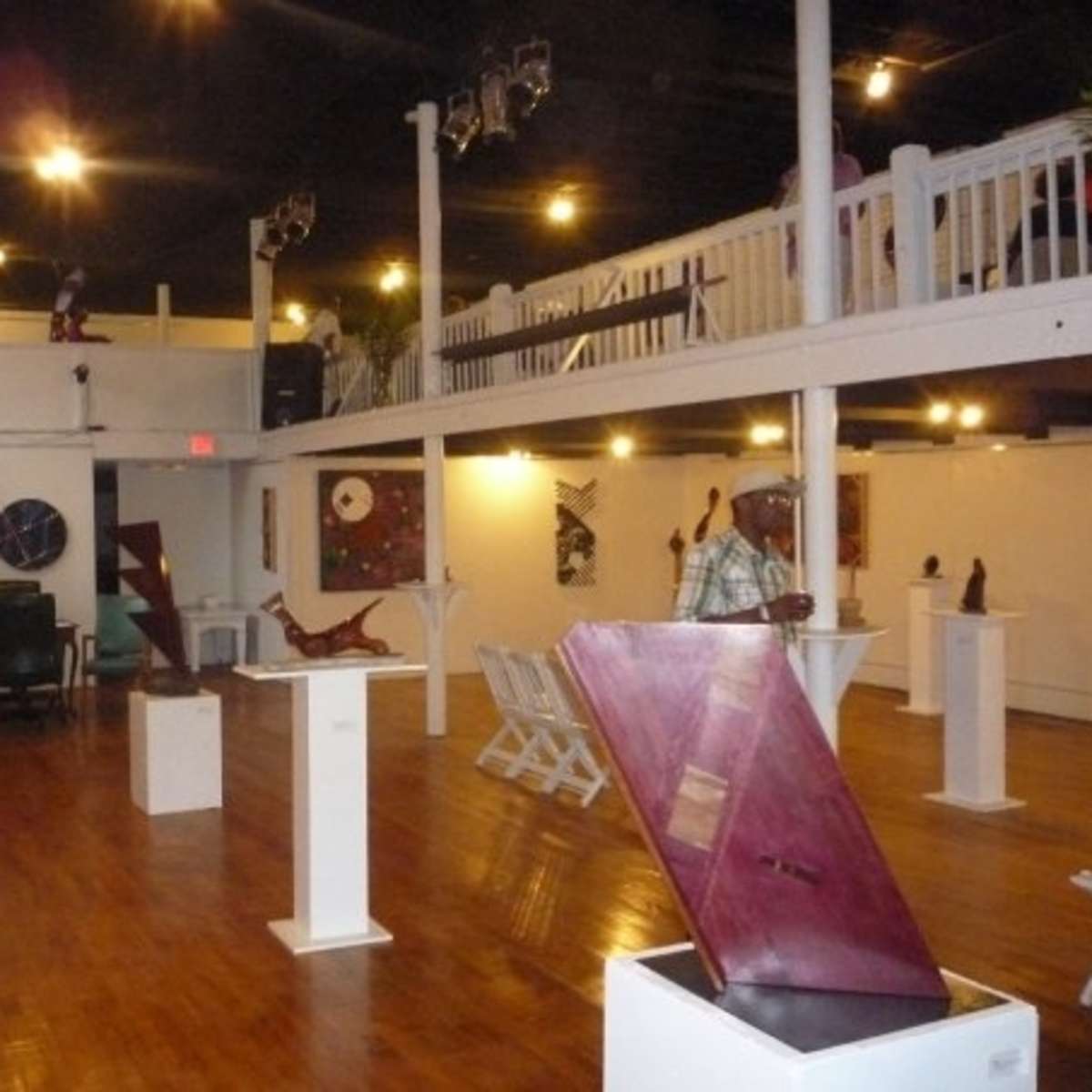 An art gallery featuring wooden sculptures displayed on pedestals, with wooden flooring and a staircase leading to a balcony above.