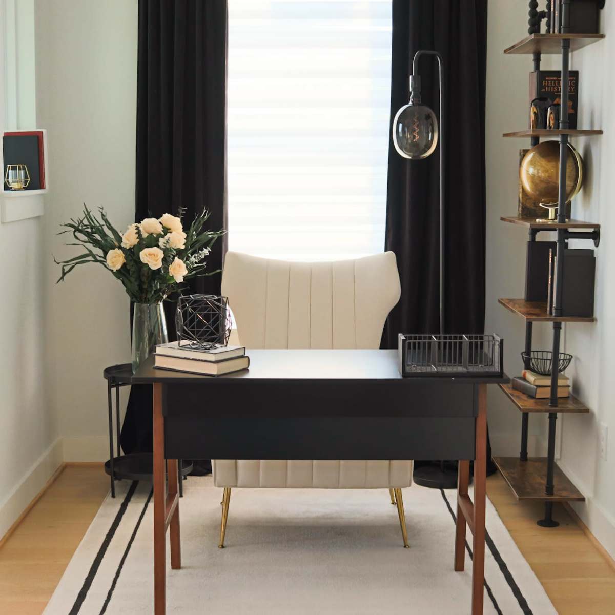 A modern office space features a black desk, a white armchair, shelving with decorative items, and a window dressed with black curtains.