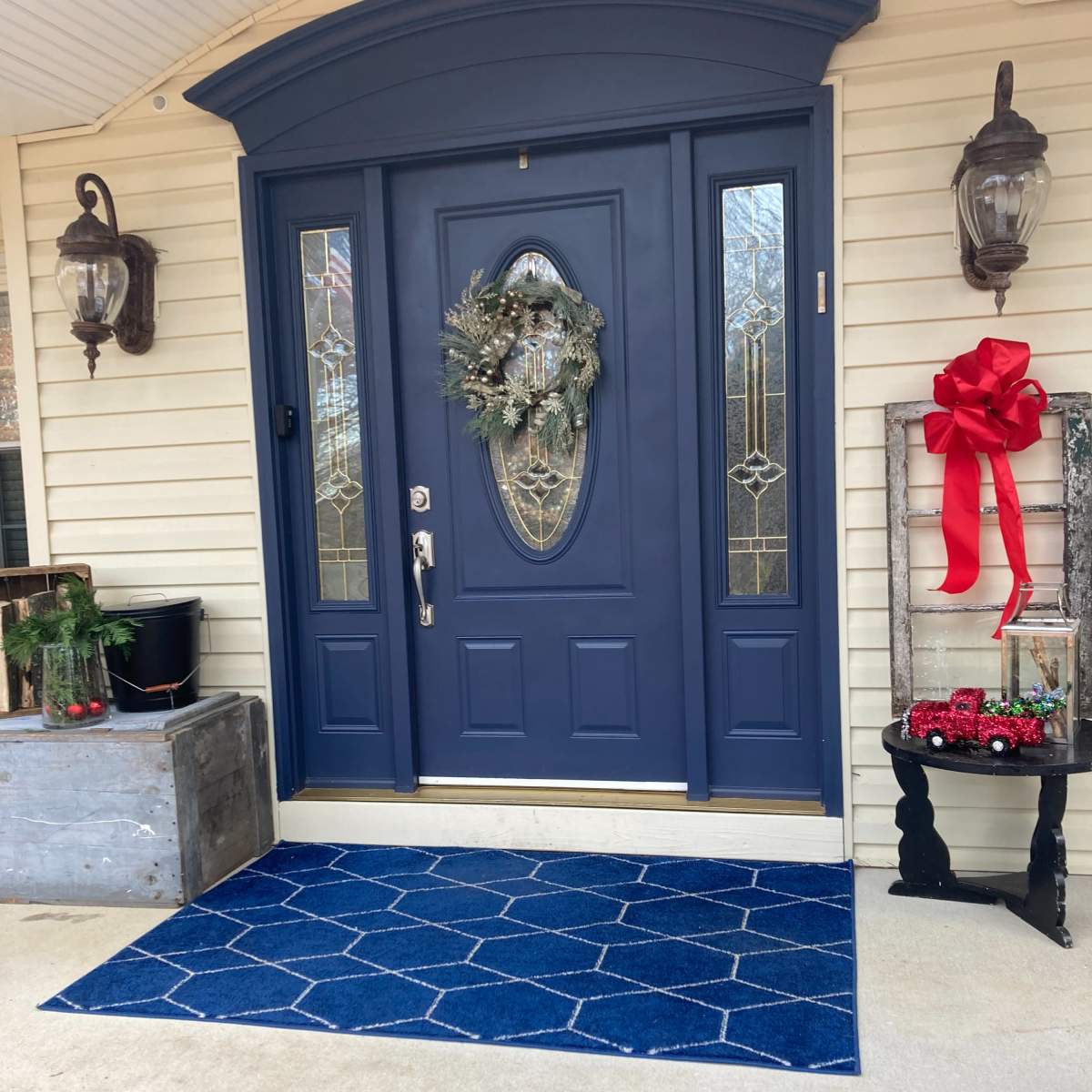A blue front door adorned with a wreath, flanked by two wall-mounted lanterns, and a decorative display featuring a red bow and greenery next to a patterned blue mat.