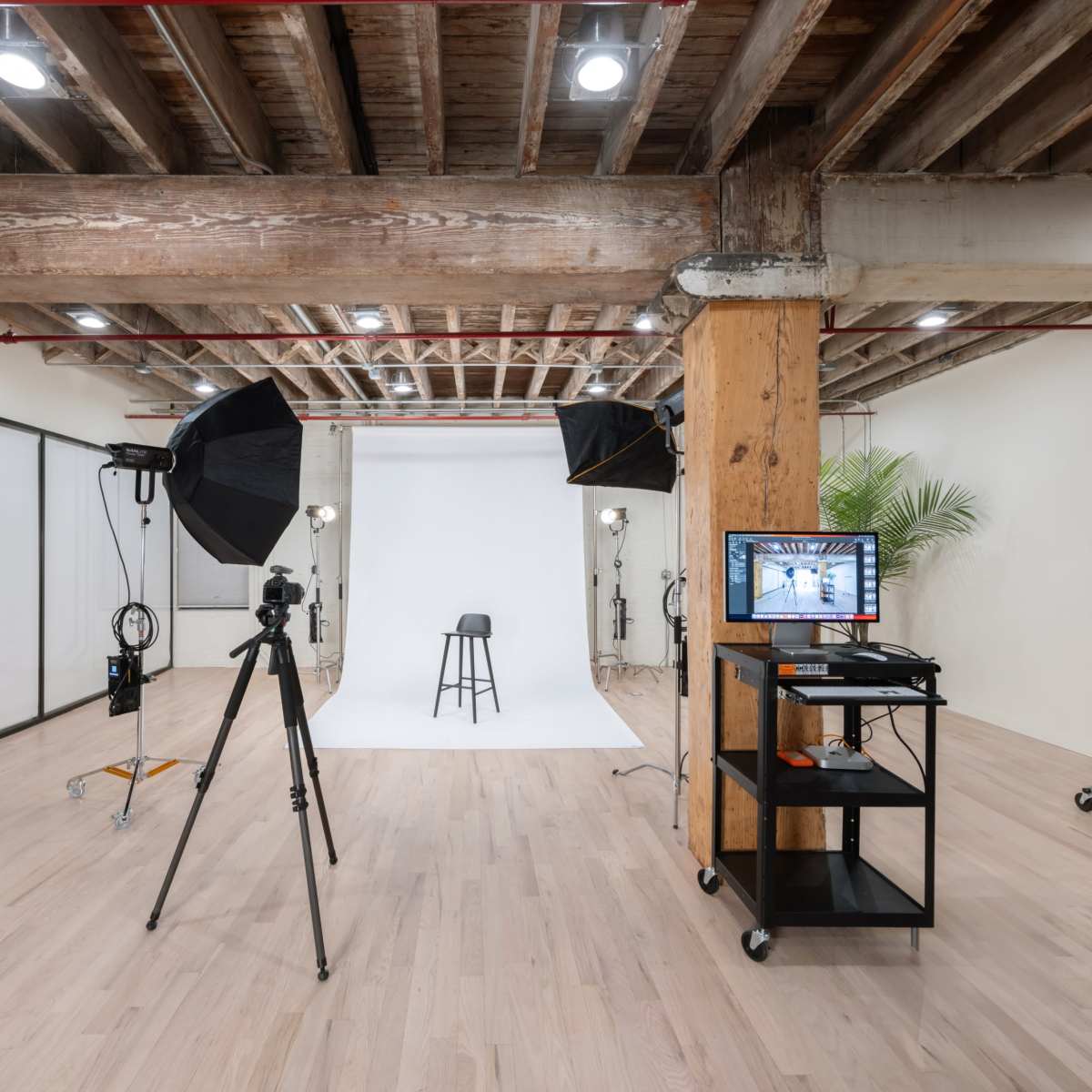 A photography studio features a white backdrop, a stool, lighting equipment, a camera on a tripod, and a monitor on a mobile cart.