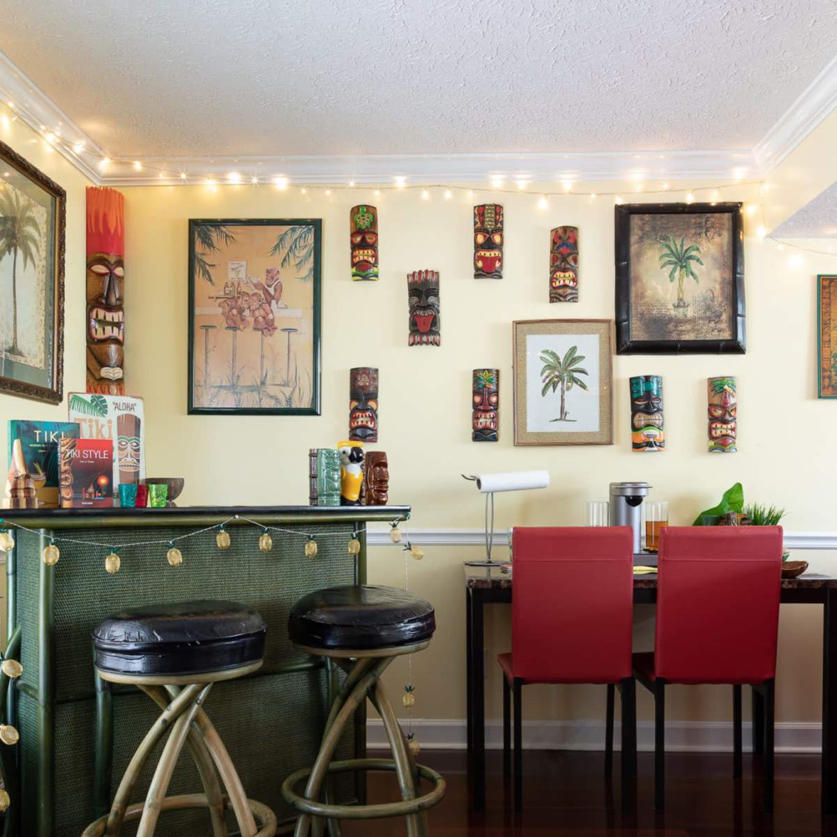 A brightly lit dining area features a bar with two black stools, a table with red chairs, and various framed artworks and tiki mugs adorning the walls.