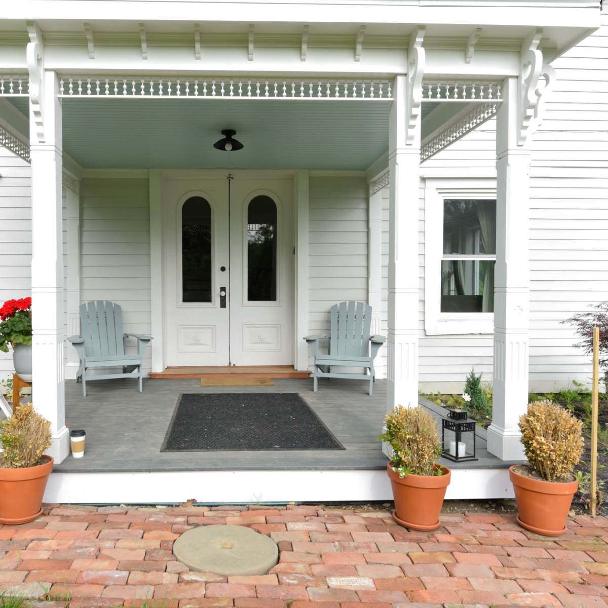 The front porch features two light blue chairs, potted plants, and a welcome mat in front of a pair of double doors.