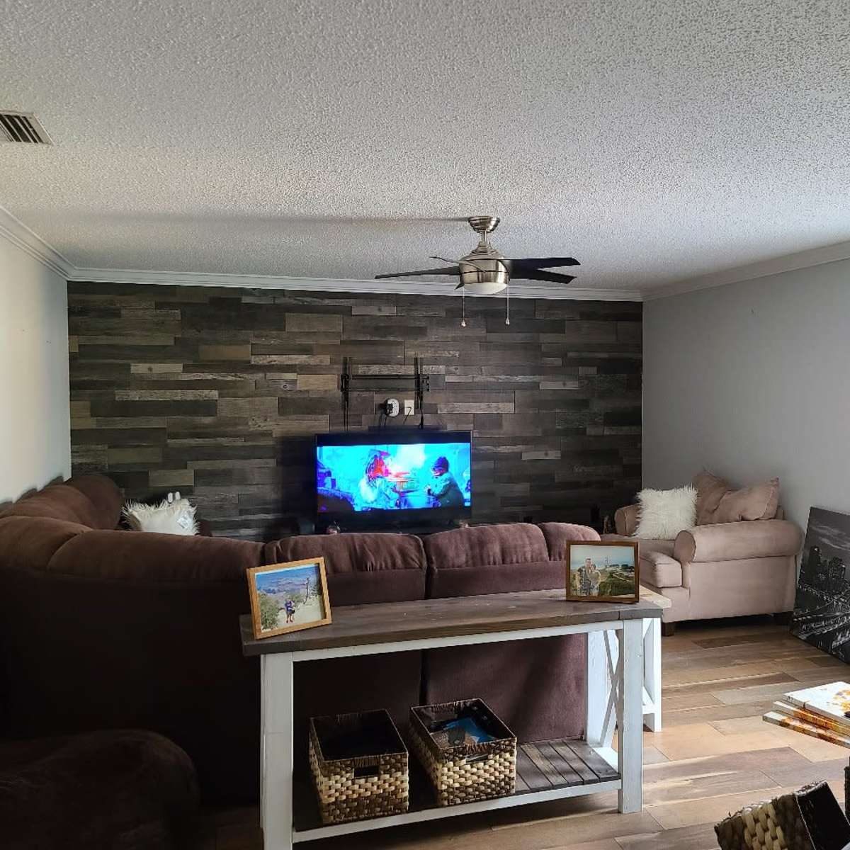 The living room features a large sectional couch facing a television mounted against a textured wall, with a coffee table in front and a casual seating area to the side.