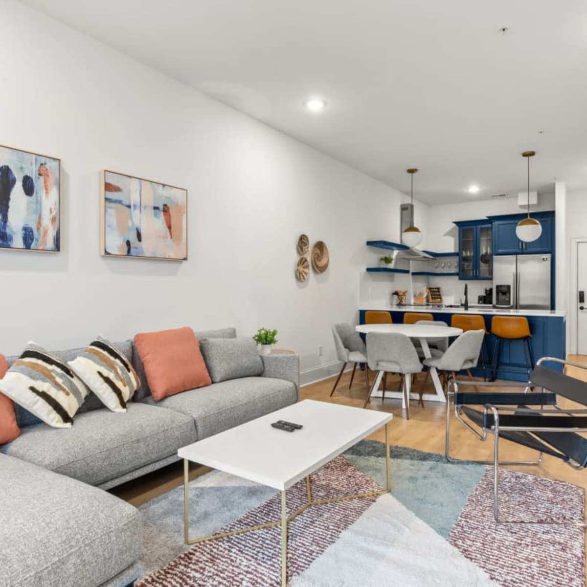 The living area features a gray sectional sofa with colorful pillows, a white coffee table, and access to a modern kitchen with blue cabinets and bar seating.